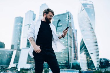 Şişkin, gözlüklü, beyaz gömlekli, kırmızı sakallı, modern şehirde cep telefonunda sörf yapan gökdelenlerle yürüyen akıllı bir erkek. 