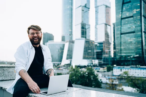 Modern dizüstü bilgisayarda yetenekli samimi hipster bir adamın portresi açık havada çalışan mutlu beyaz bir adam şehir ortamında portatif netbook 'la oturmuş ve neşeyle gülümsüyor.