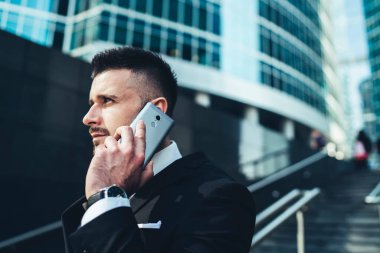 Şehir merkezindeki modern cam binalara karşı cep telefonuyla konuşan takım elbiseli genç bir adamın yan görüntüsü. 
