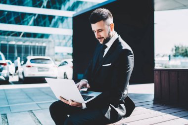 Güneşli yaz gününde iş merkezinin önündeki bankta otururken dizüstü bilgisayar ve gazete okuyan resmi siyah takım elbiseli modern yöneticinin yan görüntüsü.