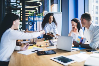 Odaklanmış Afro-Amerikalı iş kadını resmi kıyafetler giyip toplantı sırasında çalışanlarla konuşurken ofiste çeşitli aletlerle masanın yanında duruyor.