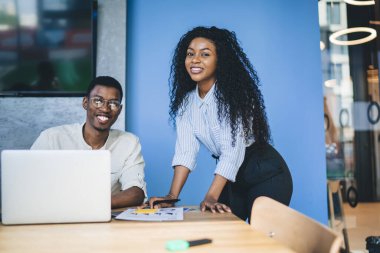 Modern resmi kıyafet giyen, dizüstü bilgisayarda sörf yapan, modern iş yerinde doküman inceleyen ve kameraya bakan Afrikalı Amerikalı ortaklar.