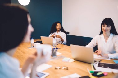 Modern ahşap konferans masasında çeşitli ofis gereçleri ve aletleriyle otururken taşınabilir bilgisayarlar üzerinde çalışan kadın ofis çalışanları.