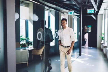 Resmi olarak kararlı Afrikalı Amerikalı bir adam elinde akıllı bir telefonla modern ofis binasının cam duvarıyla lobide geziniyor.