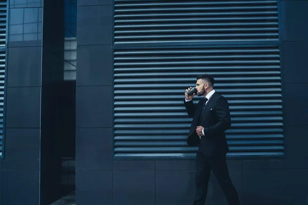 街の通りに黒い現代的な建物の外を歩きながら ジャケットや飲み物を調整ひげそりのビジネスマンの側面図 — ストック写真