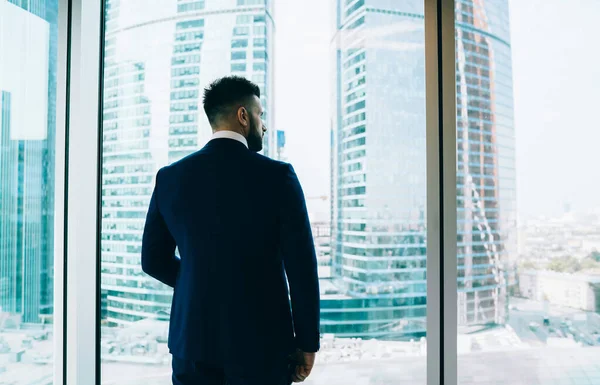 オフィスの窓の前に立つ正式なスーツ姿の認識できないマネージャーのバックビューと現代の高層ビルを離れて見る — ストック写真