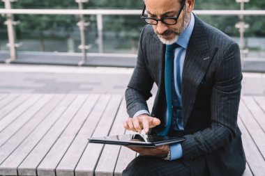 Ekin, orta yaşlı, yoğun, şık takım elbiseli bir erkek kentsel arkaplanda bankta otururken modern dijital tablet ekranına tıklıyor.
