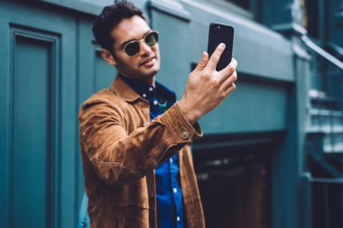 Moda elbiseli ve güneş gözlüklü genç yakışıklı New York caddesinin mavi binasında dururken akıllı telefondan selfie çekiyor.