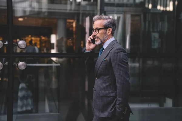 ニューヨークでポケットに手を入れてオフィスビルのガラスの壁の近くの電話で話してスタイリッシュな暗いビジネススーツで髭を生やしたビジネスマンの側面図 — ストック写真