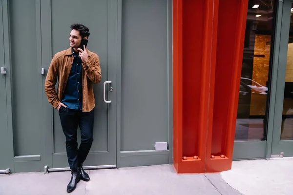 ニューヨークの通りを眺めながら電話で話す灰色の入り口にもたれて黒い髪の流行の服のハンサムな若い男性 — ストック写真