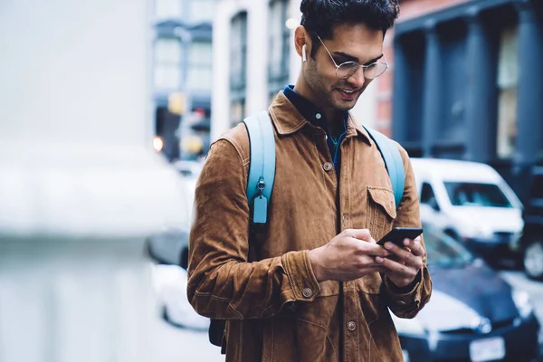 在纽约街上边走边听音乐时 身穿褐色羊皮夹克 背着背包和耳机用手机发短信的英俊的西班牙裔快乐男性戴着眼镜 — 图库照片