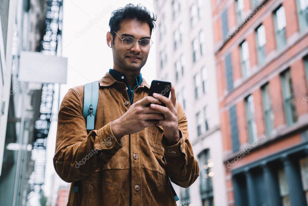 From below handsome delighted Hispanic male in eyeglasses wearing brown suede jacket with backpack and earphones standing on sidewalk in New York city and typing on cellphone