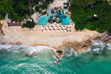 Açık yüzme havuzu ve güneş pansiyonları olan deniz manzaralı otel oteli. Yaz tatilinin kuş bakışı manzarası, turkuaz deniz suyu ve temiz kum kıyısındaki şemsiyeler