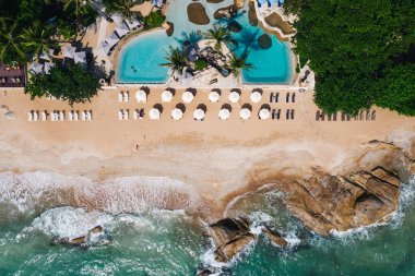 Açık yüzme havuzu ve güneş pansiyonları olan deniz manzaralı otel oteli. Yaz tatilinin kuş bakışı manzarası, turkuaz deniz suyu ve temiz kum kıyısındaki şemsiyeler