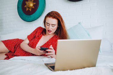 Konsantre olmuş, ağzı açık, benekli, dizüstü bilgisayarın yanında yatıp evde akıllı telefondan mesaj atan kızıl saçlı kadın.