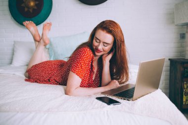 Utangaç, kızıl saçlı, parlak noktalı elbiseli bir kadın evde akıllı telefon ve dizüstü bilgisayar kullanırken yumuşak yatağa uzanıyor.