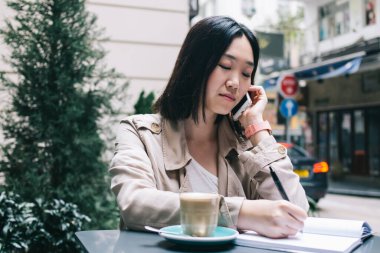 Konsantre olmuş, kısa siyah saçlı, elinde kahve fincanı ve akıllı telefonuyla masada oturan şehir yolunun arka planına karşı deftere yazı yazarken konuşan genç bir bayan.