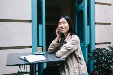 Kısa siyah saçlı, elinde kahve ve defterle masada oturan ve akıllı telefonuyla konuşan genç bir kadının yan görüntüsü.