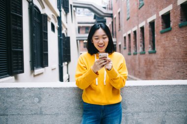 Sarı kazaklı genç Asyalı kadın taş çitlere yaslanıyor ve şehrin arka planındaki binalara karşı akıllı telefon kullanarak gülümsüyor.