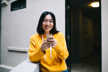 Koyu renk kısa saçlı, sarı kapüşonlu, elinde telefonla kameraya gülümseyen genç bir Asyalı kadın.