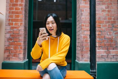 Genç, kısa saçlı, sarı kapüşonlu ve yırtık pantolonlu bir kadın. Bankta oturmuş, ağzı açık bir şekilde ekrana bakıyor.