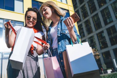 Aşağıdaki meraklı, kaygısız kadınların alışveriş merkezinde yürürken giysi almalarından sonra zevkle alışveriş çantalarına bakmaları.
