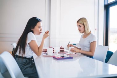 Beyaz tişörtlü çok ırklı kadınlar masada oturup kahve içip, modern beyaz kafeteryada kek tadıyorlar. 