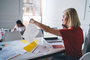 Cep telefonuyla konuşan, elinde belge tutan, ofisteki bilgisayarı açık bir masada bilgi arayan düşünceli, kendine güvenen bir kadının yan görüntüsü.