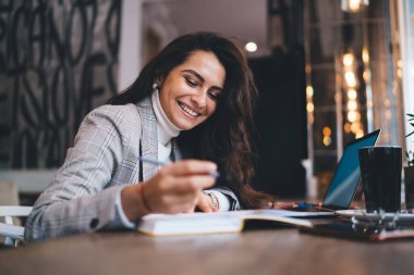 Uzun siyah saçlı, sıradan giyinmiş, zeki ve neşeli bir bayan. Kafede dizüstü bilgisayarla oturup notlar alıyor.  