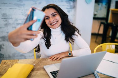 Çalışırken ve kafeteryada dizüstü bilgisayar ve not defterlerinin önünde otururken mutlu bir kadın cep telefonuyla selfie çekiyor.