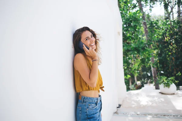 Wanita Keriting Berbicara Telepon Genggam Dengan Senyum Menawan Bersandar Dinding — Stok Foto