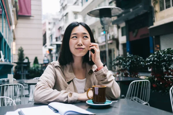 穿着休闲装的快乐的亚洲女人一边坐在餐桌旁一边 一边用智能手机说话 一边在咖啡店里望着远方 — 图库照片