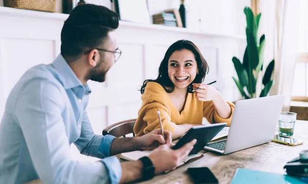 Pleased woman and male in glasses wearing casual clothes writing notes in notebooks while browsing devices and sitting at table