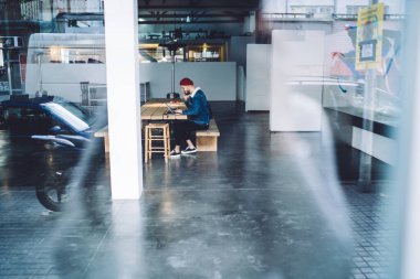 Kırmızı beany ve kot ceketli profesyonel yetişkin işçinin yan görüntüsü ahşap masada oturmuş laptopta oturmuş modern ofiste telefonla konuşuyor.