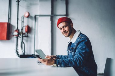 Sıcak mavi kot ceketli ve kırmızı şapkalı neşeli, genç, şık, sakallı, serbest çalışan, iş yerinde telefon kullanan, kameraya eğilmiş, 