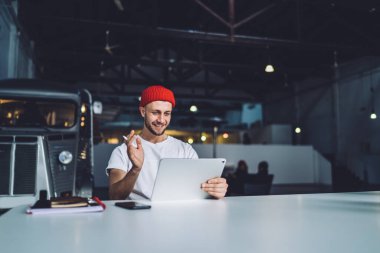 Kırmızı bereli genç adam büyük tablet seyrediyor. Stylus kalemle çalışıyor ve garajda çalışırken mutlu bir şekilde gülümsüyor. 