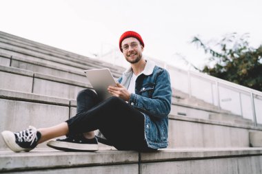 Gözlüklü, kırmızı şapkalı ve kot ceketli gülümseyen sakallı genç adam beton bankta oturuyor ve açık havada tabletle birlikte arka plandaki bulanık kameraya bakıyor.