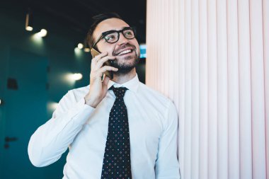 Gözlüklü, beyaz gömlekli, kravatlı, cep telefonuyla konuşan, beyaz duvara yaslanan neşeli mutlu adam.  