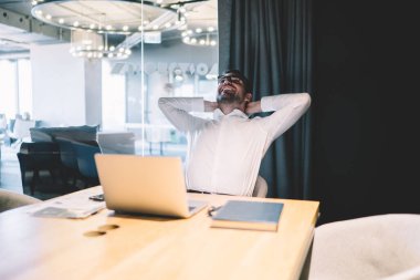 Resmi tişörtlü, elleri başının arkasında olan neşeli bir adam. İşyerindeki şirketin başarısını kutlayan projenin sonuçlarını kontrol etmek için laptop kullanıyor.
