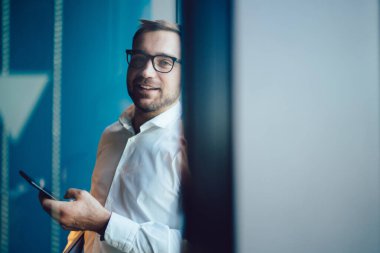 Optik gözlüklü, kendine güvenen bir adamın yarım boy portresi iş arasında kameraya bakan, modern akıllı telefon cihazlı yakışıklı beyaz erkek yönetmen.