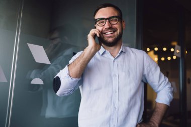 Mavi gömlekli ve mavi pantolonlu neşeli adam ofisin cam kapısına yansıyan cep telefonuyla sohbet ediyor.