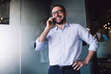Ofiste online telefon görüşmeleri sırasında akıllı telefon iletişiminden keyif alan, optik gözlüklü neşeli bir erkek bankacı finans müdürüyle konuşuyor ve gülüyor.