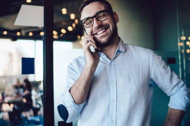 Mavi gömlekli, gülen beyaz dişli adam cam kapının karşısındaki ofiste cep telefonuyla konuşuyor.