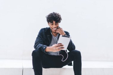 Afro-Amerikan gülümseyen genç erkek çenesine dokunuyor ve beyaz merdivenlerde otururken tablette selfie çekiyor ve kulaklıkla beyaz duvara karşı müzik dinliyor.