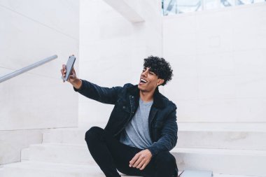 Şen şakrak, kıvırcık, kotlu bir adam. Akıllı telefonuyla beyaz merdivenlerde oturuyor. Parlak bir odada, selfie çekiyor ve gülümsüyor.