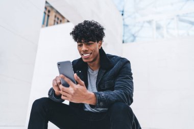 Kıvırcık saçlı, şık giyinmiş, beyaz, modern bir binada otururken akıllı telefon kullanan, gülümseyen, Afro-Amerikan bir adam.