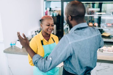 Önlüklü, önlüklü, birbirleriyle sohbet eden Afro-Amerikalı genç ve modern meslektaşlar şehirdeki kafeteryada keklerle tezgahta duruyorlar.