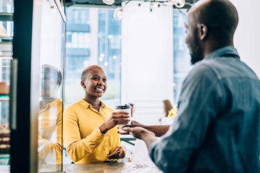 Sarı tişörtlü, gülümseyen, siyah bir bayan, şehirdeki kafede çalışan Afrikalı Amerikalı bir çalışanın kahvesini içiyor.
