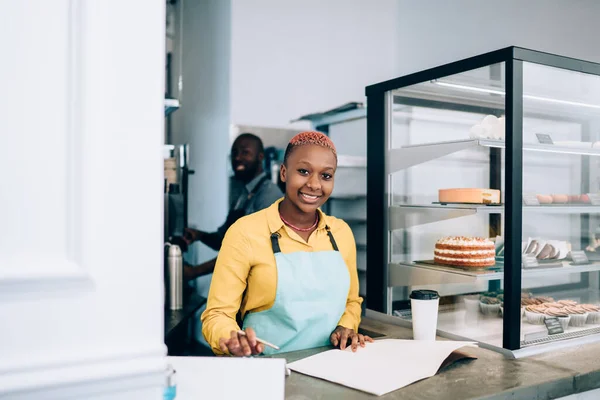 快乐可爱的非洲裔美国女士在围裙上写下各种甜点 与咖啡店里笑着的黑人合作 看着镜头 — 图库照片