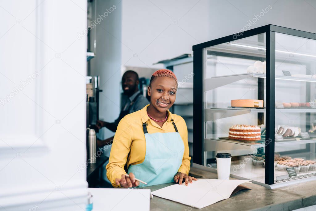 Cheerful lovely African American lady in apron writing down dessert assortment working with black smiling guy in cafe looking at camera